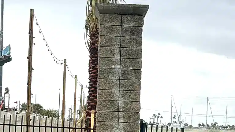 420government-stonecolumn Tall rectangular stone column beside a waterfront walkway, with string lights, palm trees, marina masts, and an overcast sky in the background.