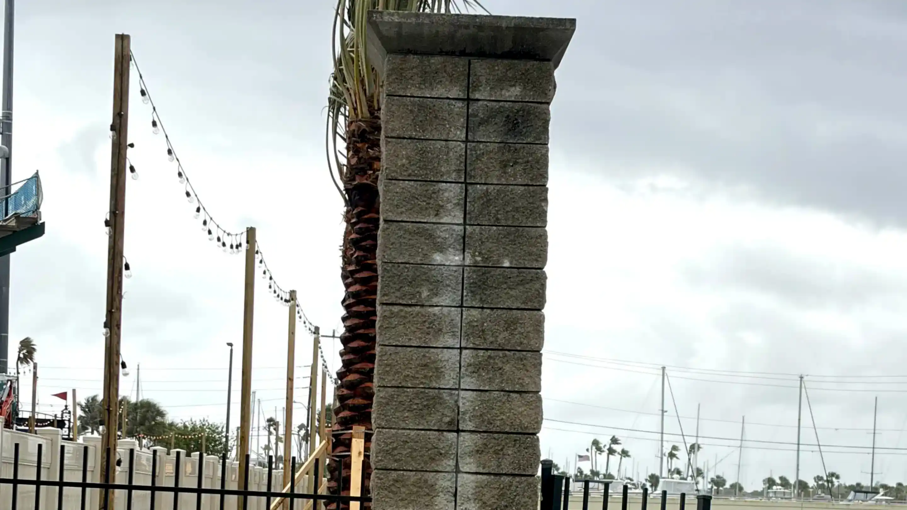 Tall rectangular stone column beside a waterfront walkway, with string lights, palm trees, marina masts, and an overcast sky in the background.