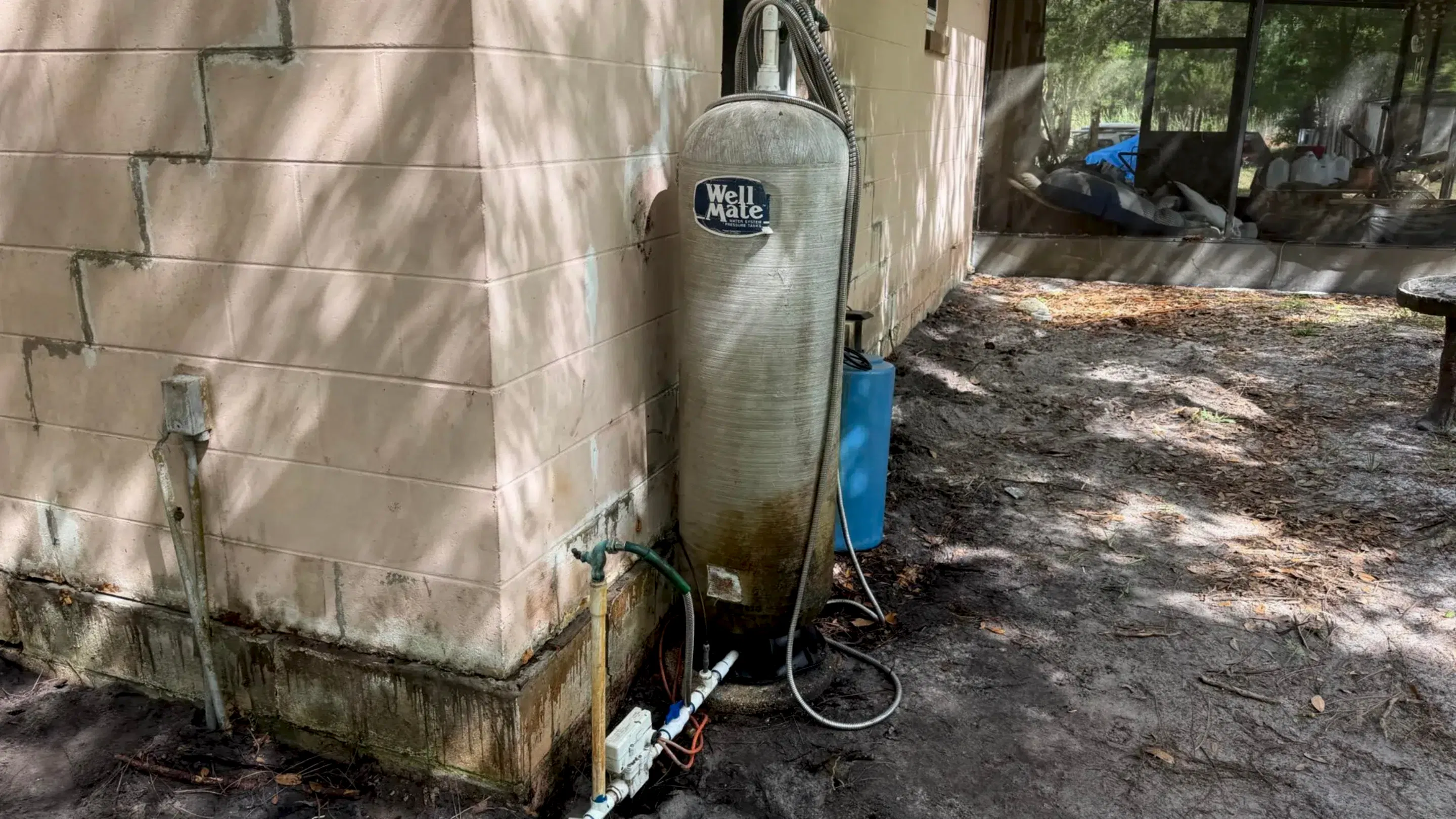 Exterior well system with a large pressure tank, PVC piping, and moisture staining along the block wall.