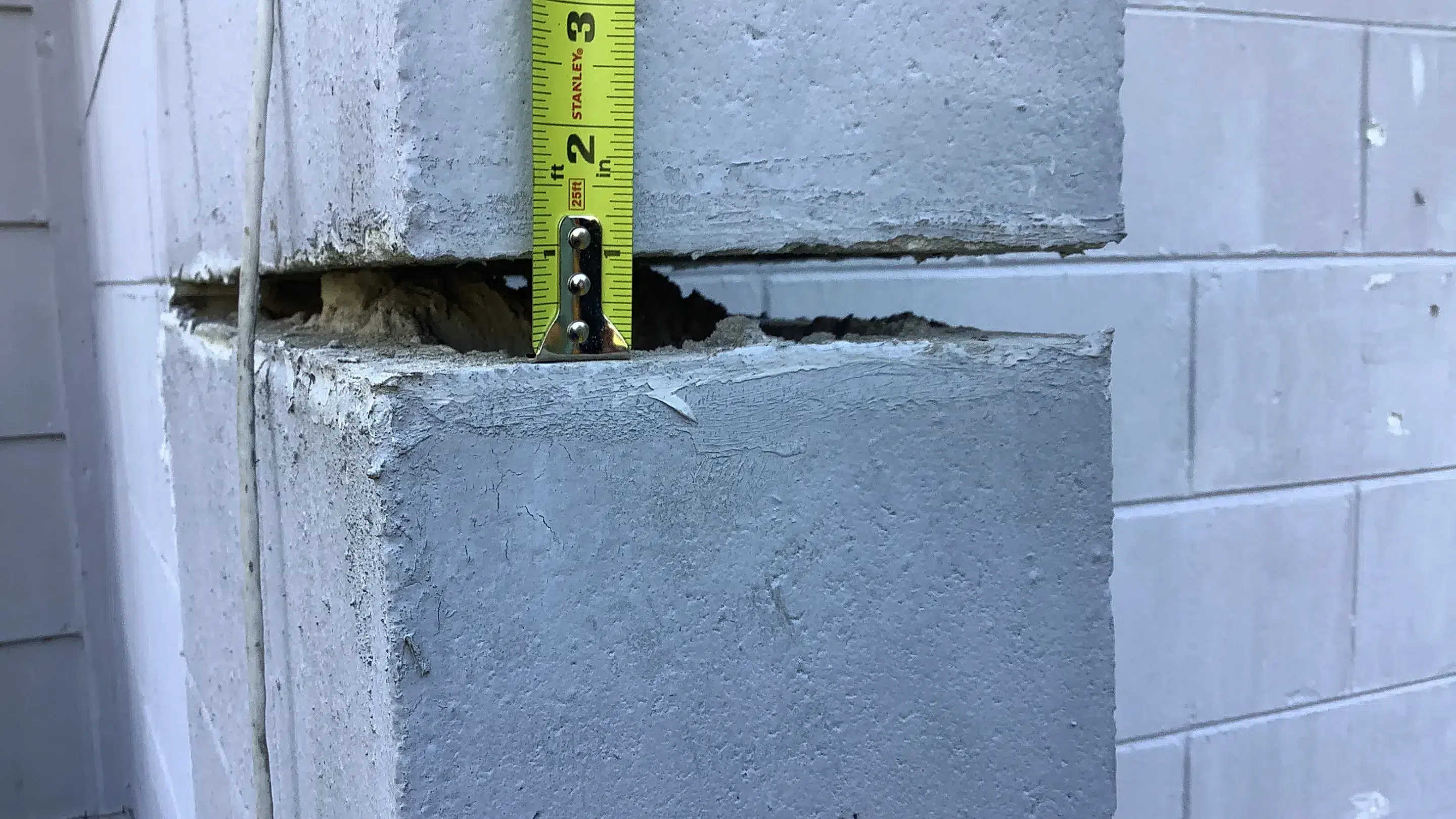Measuring the width of a horizontal gap between displaced concrete block sections in a residential foundation wall using a tape measure to document structural separation.